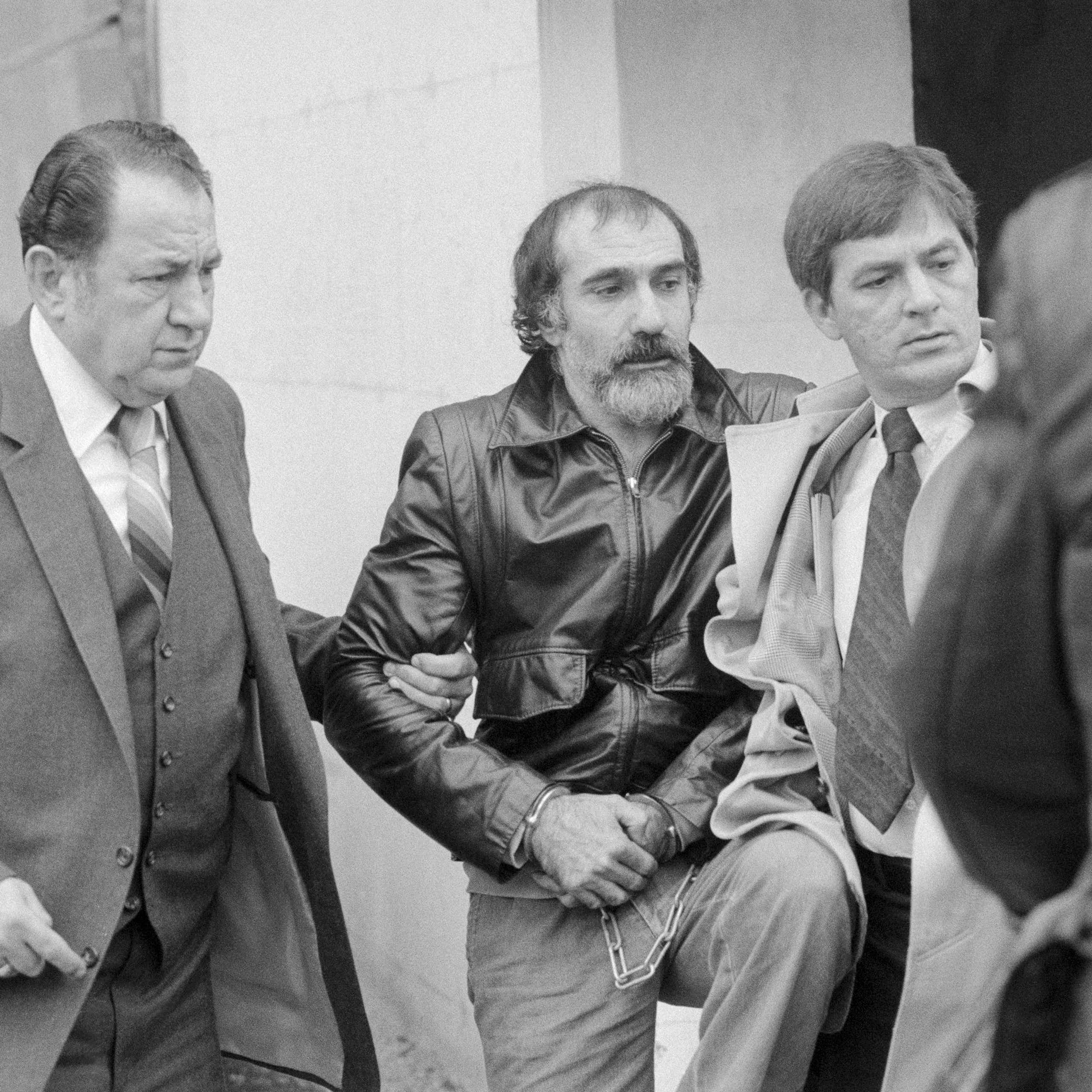 Black and white photo of men escorting handcuffed man with mustache and beard and wearing a leather jacket