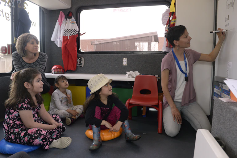 An adult kneels and write on a whiteboard in black marker as children sit on pads and take in a lesson. Another teacher looks onward.