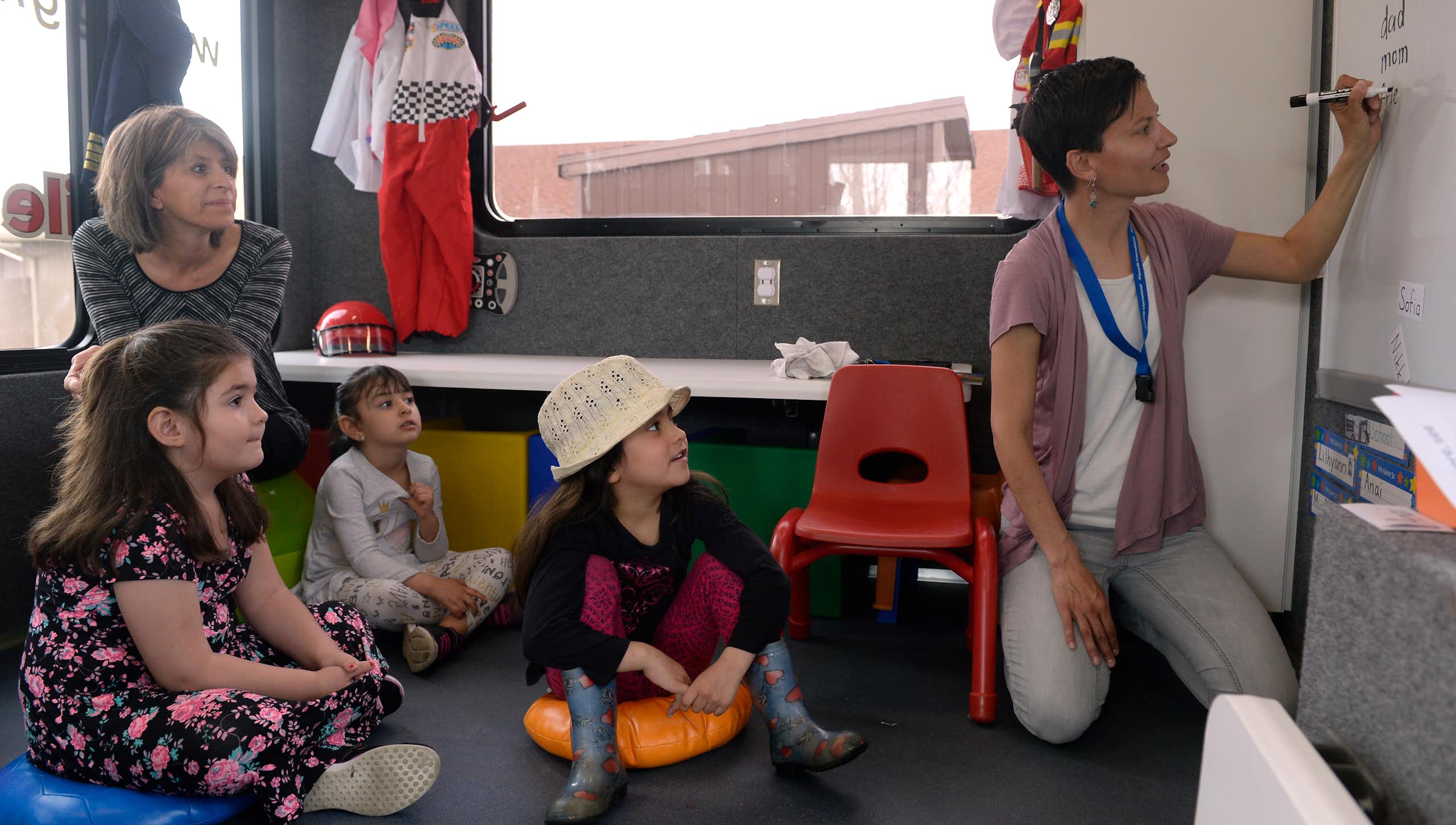 An adult kneels and write on a whiteboard in black marker as children sit on pads and take in a lesson. Another teacher looks onward.