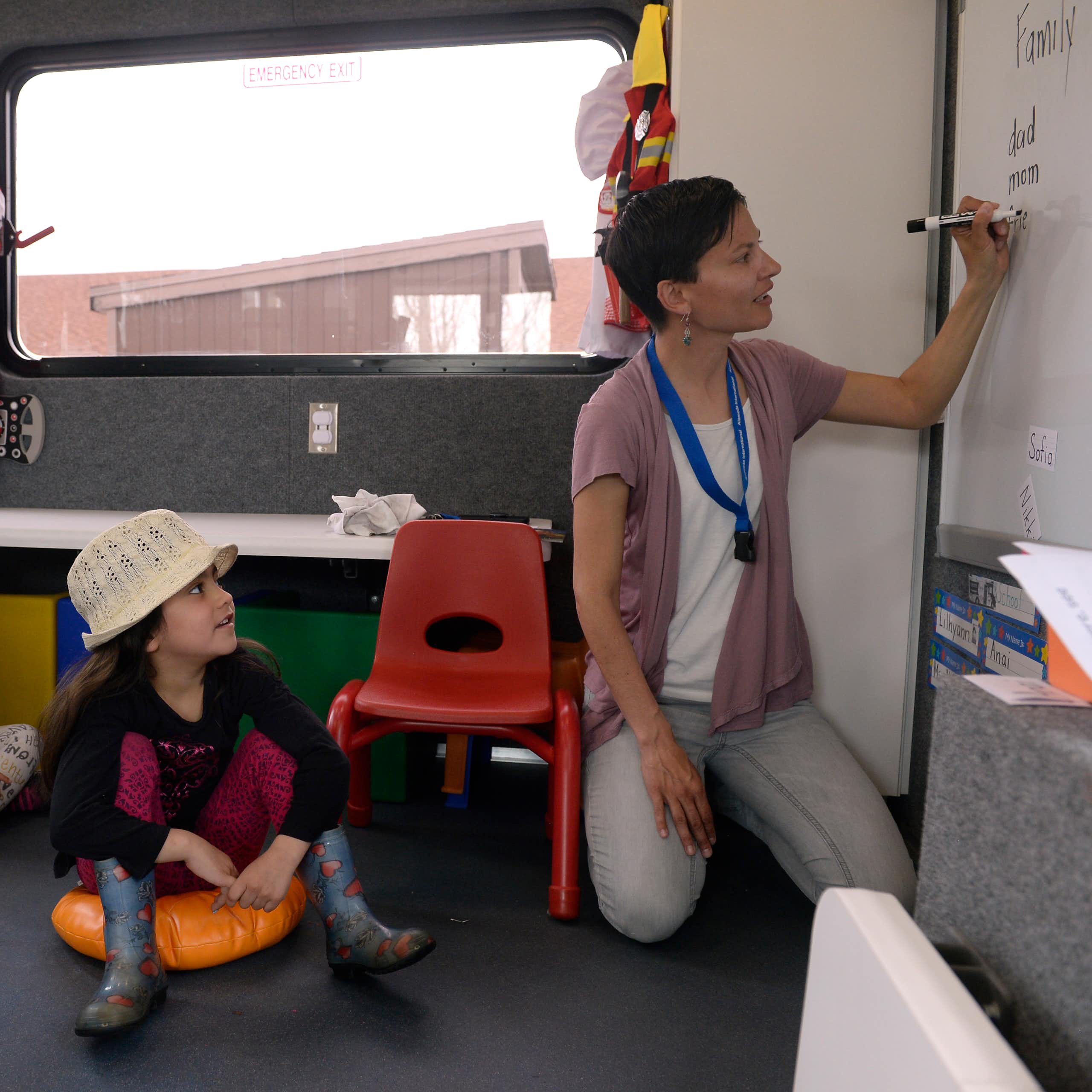 An adult kneels and write on a whiteboard in black marker as children sit on pads and take in a lesson. Another teacher looks onward.