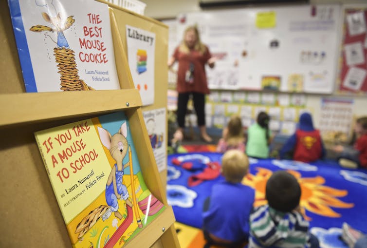 Books are in focus in the foreground with titles like 'The Best Mouse Cookie' and 'If you take a mouse to school.' In the blurred background is an adult woman standing and teaching to a bunch of young children sitting on the ground.