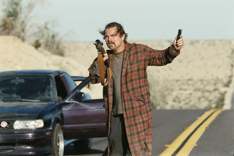 Leonardo DiCaprio holds guns on a desert road.