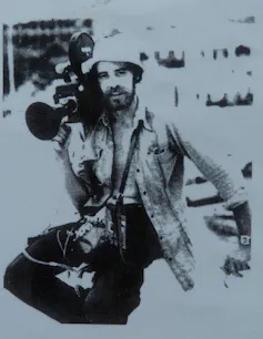 A black and white image of a man with his shirt undone, carrying a film camera on his shoulder