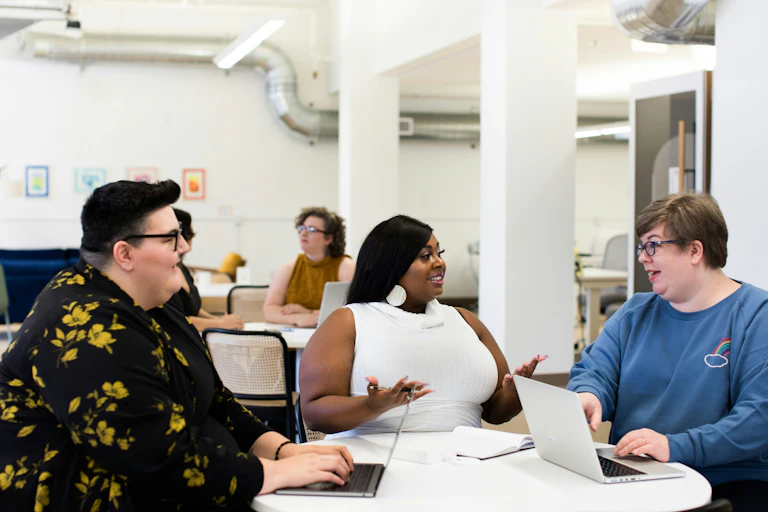Three larger-bodied people talk around a table, two of whom are using laptops