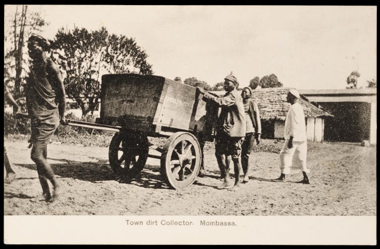 Uma foto antiga de homens africanos trabalhando. Um puxa um carrinho de madeira enquanto outros parecem carregar coisas nele.