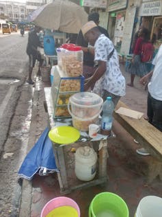 Uma cena de rua movimentada com um vendedor em uma barraca com comida exposta em recipientes e baldes de líquido ao lado dele.