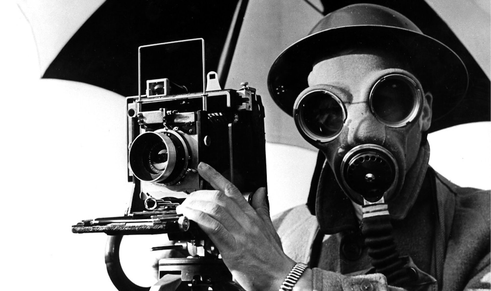 A black and white image of a photographer in a gas mask with his camera beneath an umbrella.