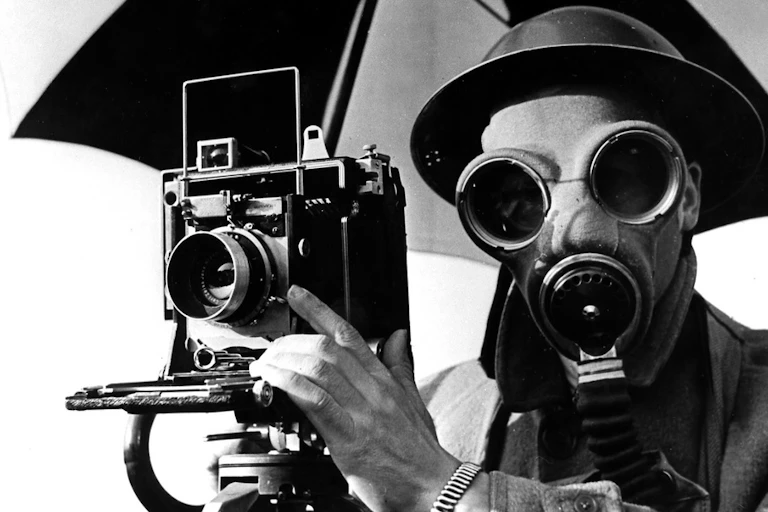 A black and white image of a photographer in a gas mask with his camera beneath an umbrella.