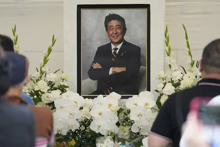 A portrait of an Asian man behind banks of white flowers with people in the foreground.