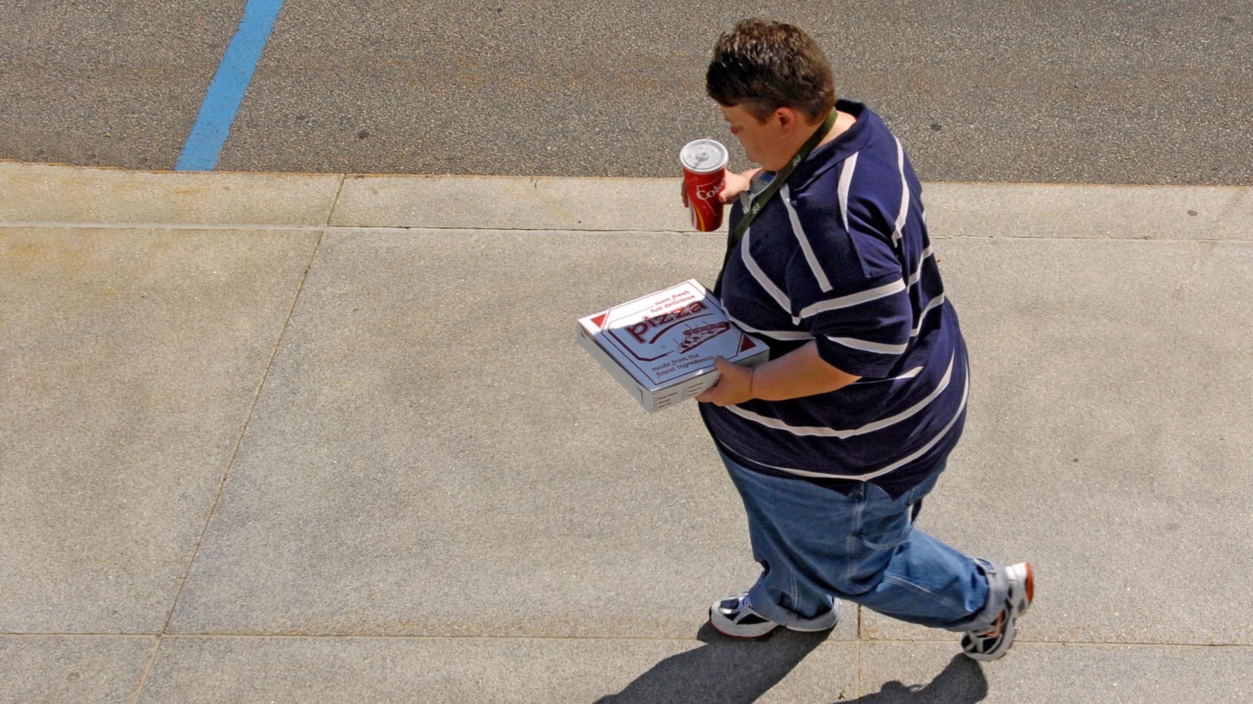 Un homme en surpoids marche dans la rue, en tenant un soda et un emballage de pizza à la main.