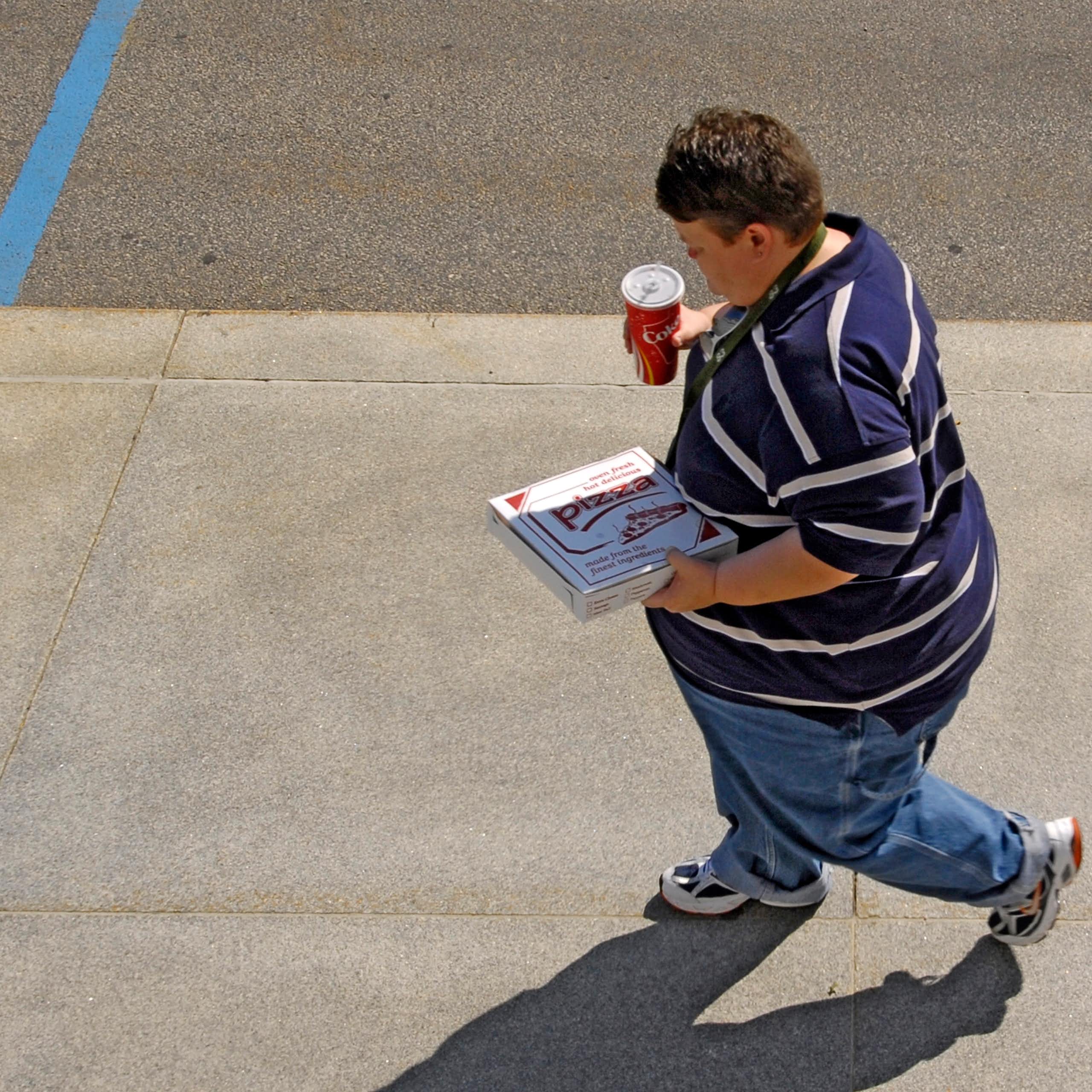 Un homme en surpoids marche dans la rue, en tenant un soda et un emballage de pizza à la main.