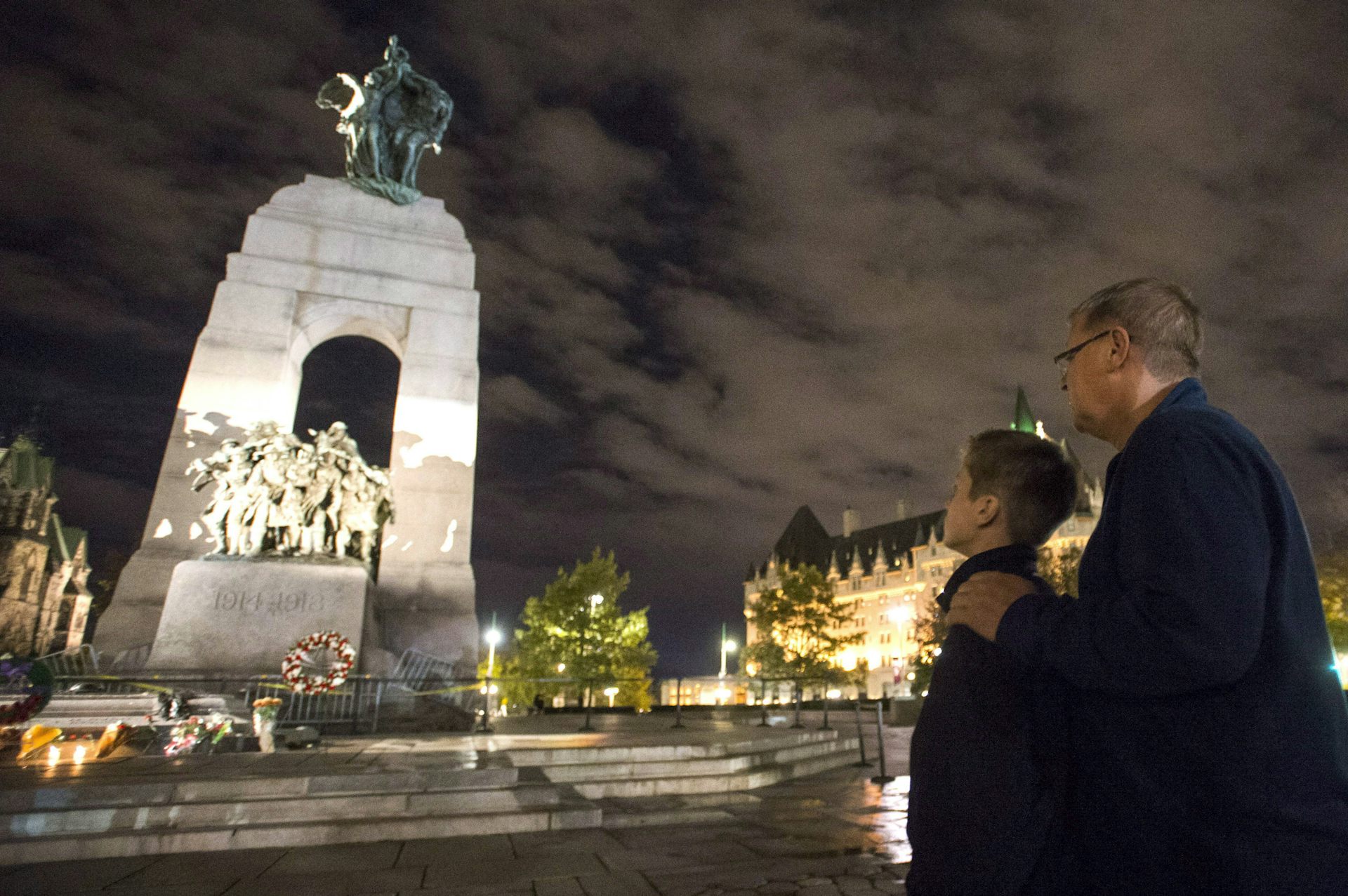 A father and son stand looking at an illuminated war memorial at dusk.