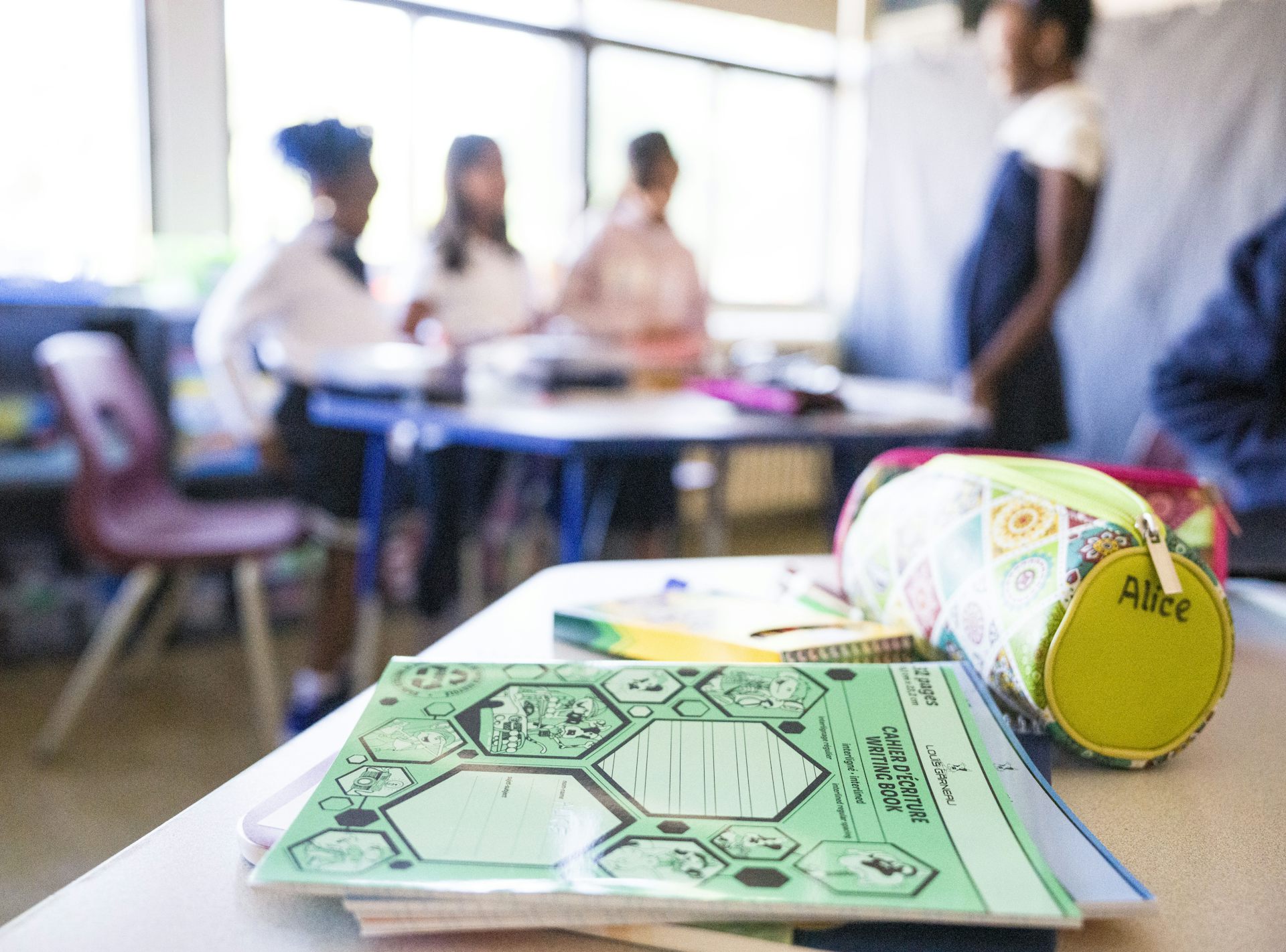 A notebook and pencil case rests on a desk with students in the background.