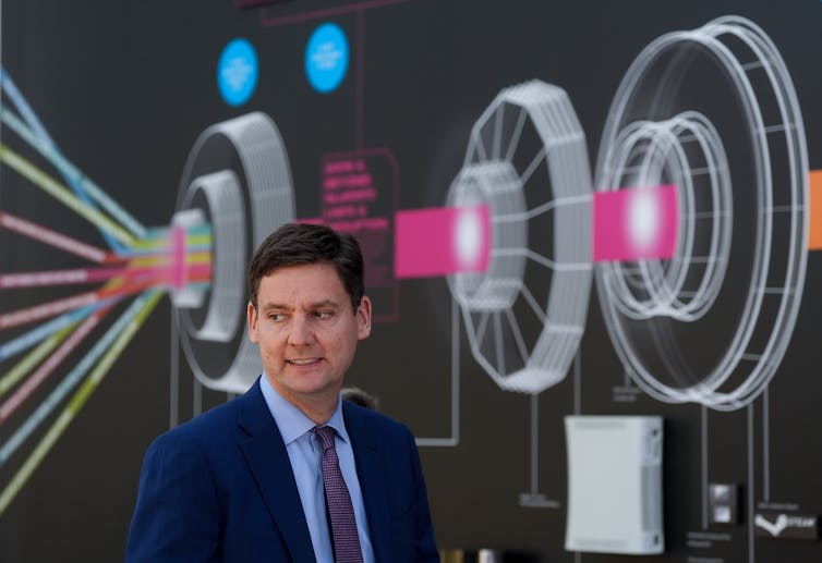 Un hombre blanco con traje se para frente a una pared decorada con imágenes de ruedas y tonos de color.