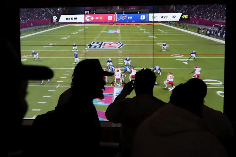 People stand in front of a large screen displaying a football video game