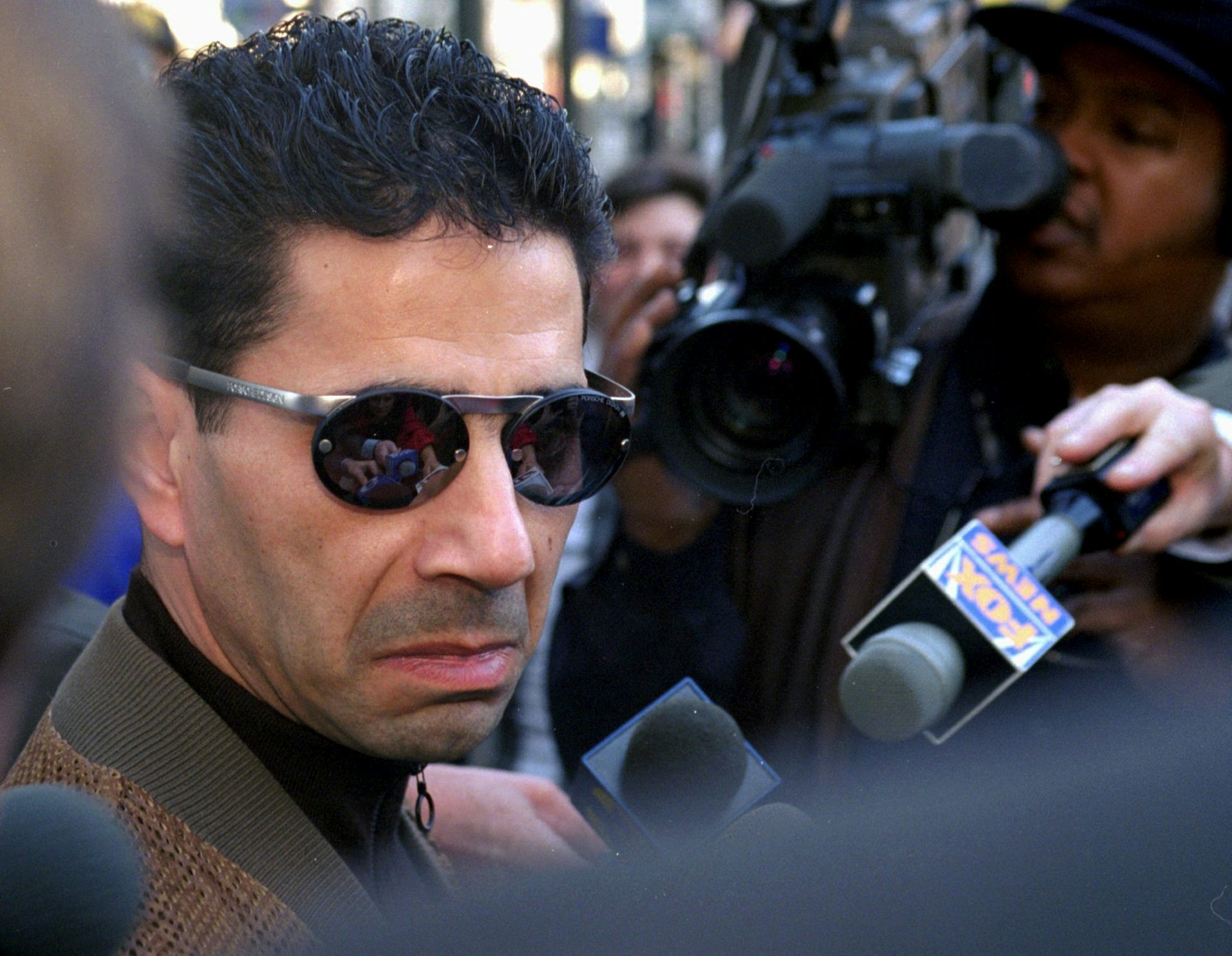 Man wearing sunglasses stands in front of people holding video camera and microphones