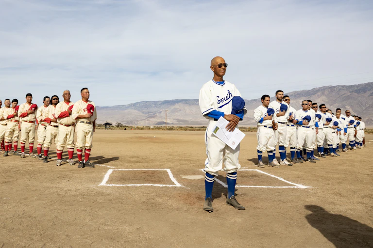 Two teams of Japanese American baseball players wearing vintage uniforms line up along the foul lines of a dusty diamond.