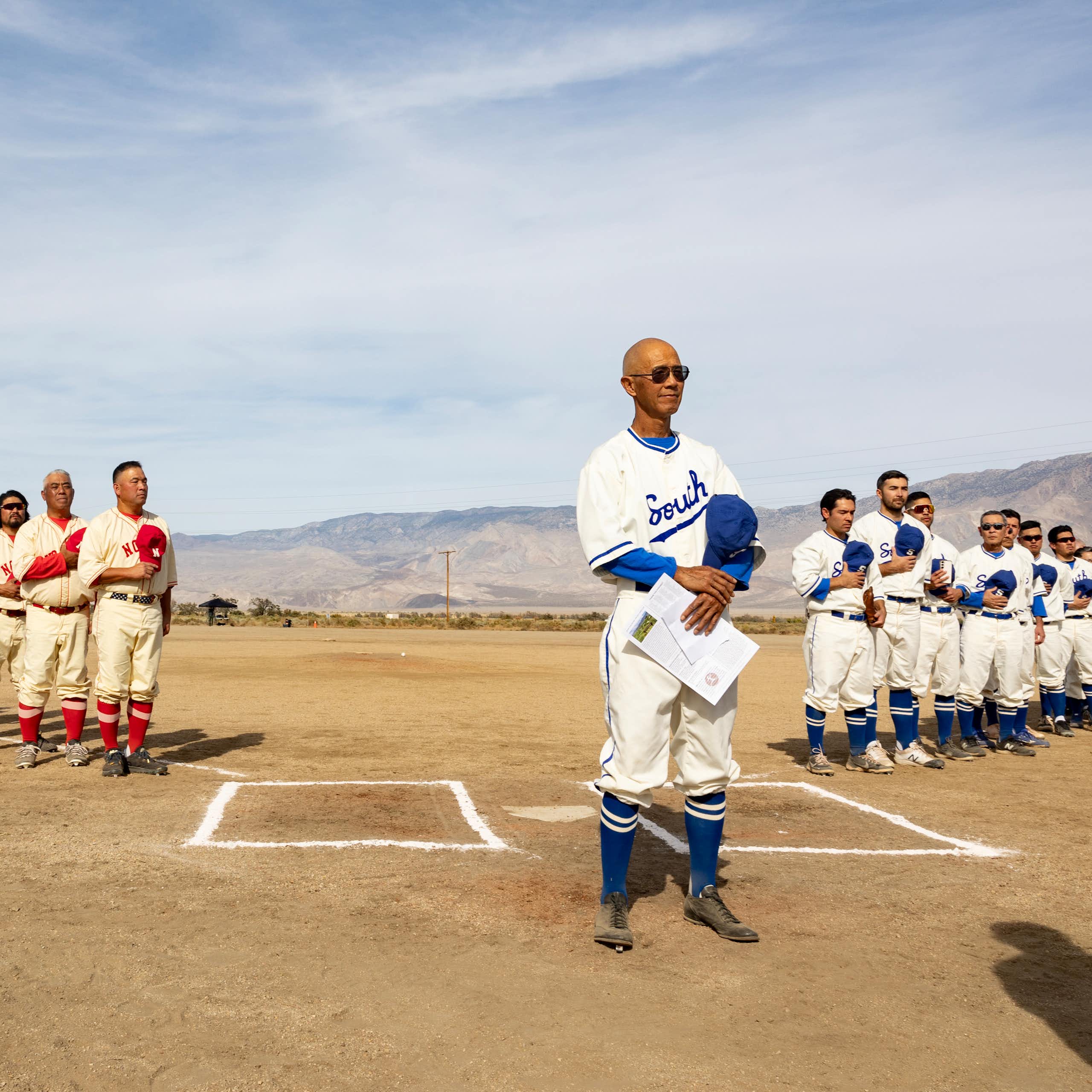 Two teams of Japanese American baseball players wearing vintage uniforms line up along the foul lines of a dusty diamond.