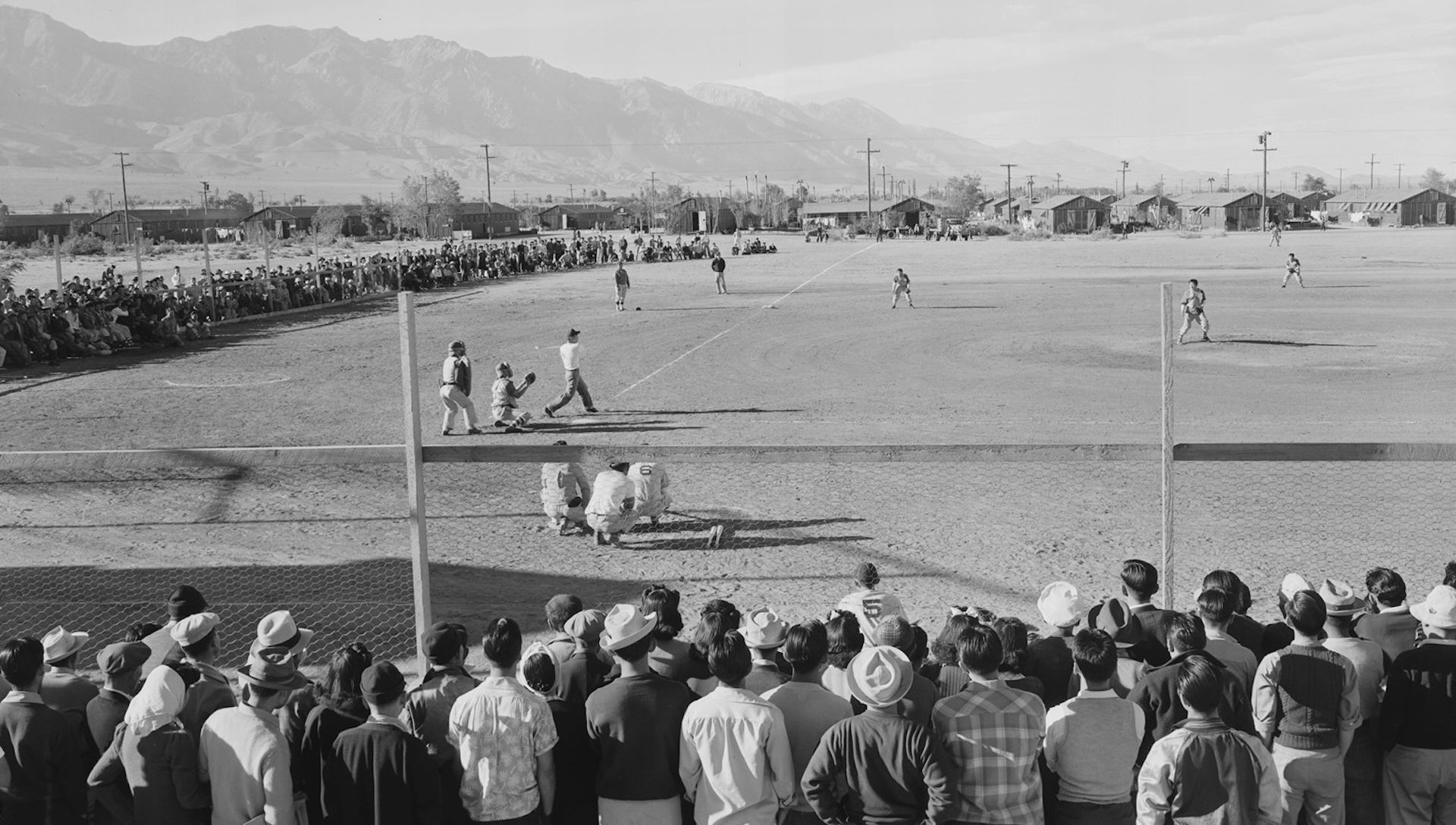 Crowds of onlookers watch a baseball game on a dusty field.