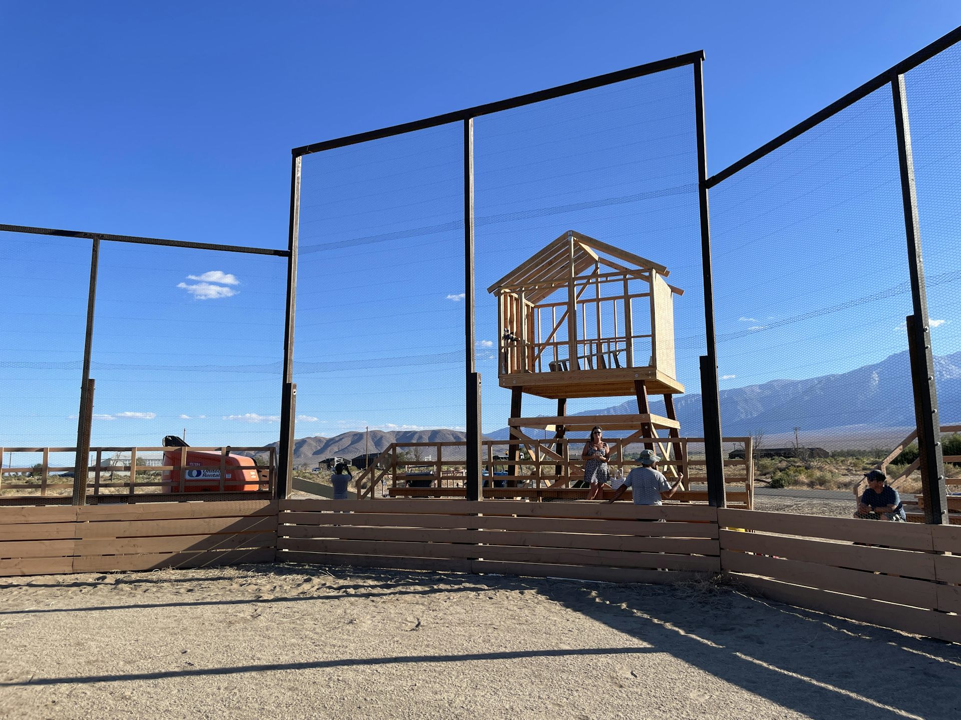 A wood-framed, enclosed booth on wooden legs behind a baseball field backstop.