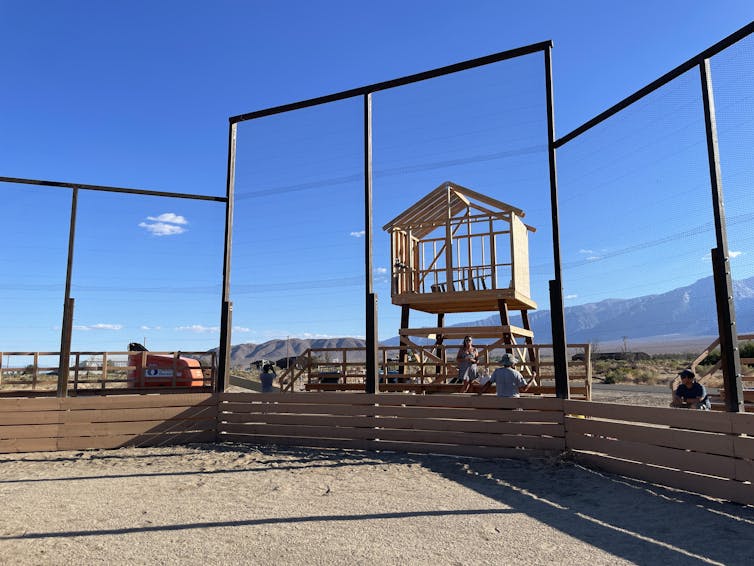 A wood-framed, enclosed booth on wooden legs behind a baseball field backstop.