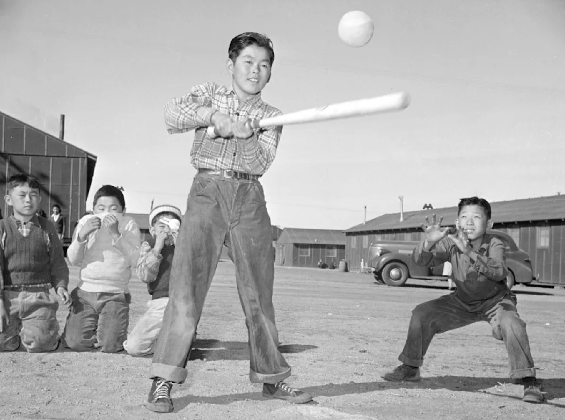An Asian American boy swings a baseball bat at an approach ball as other boys watch in the background.