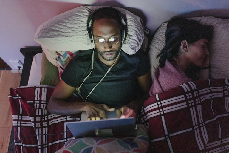 Worried man lies in bed looking at laptop while his partner sleeps.