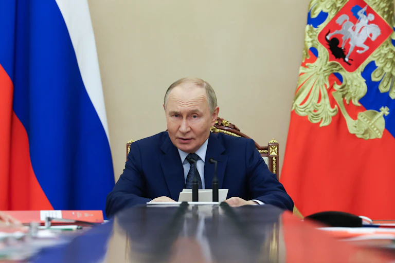 Russian president Vladmir Putin sitting at his conference table in front of national flags.