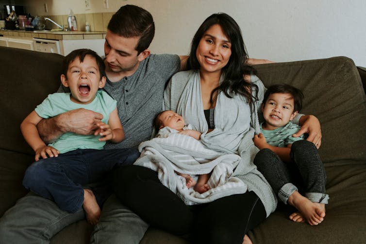 Una familia formada por un hombre, una mujer y sus tres hijos está sentada en el sofá y ríe.