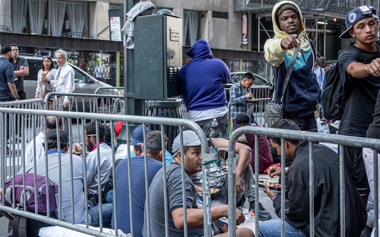 Hombres y mujeres sentados en la calle entre vallas comen.