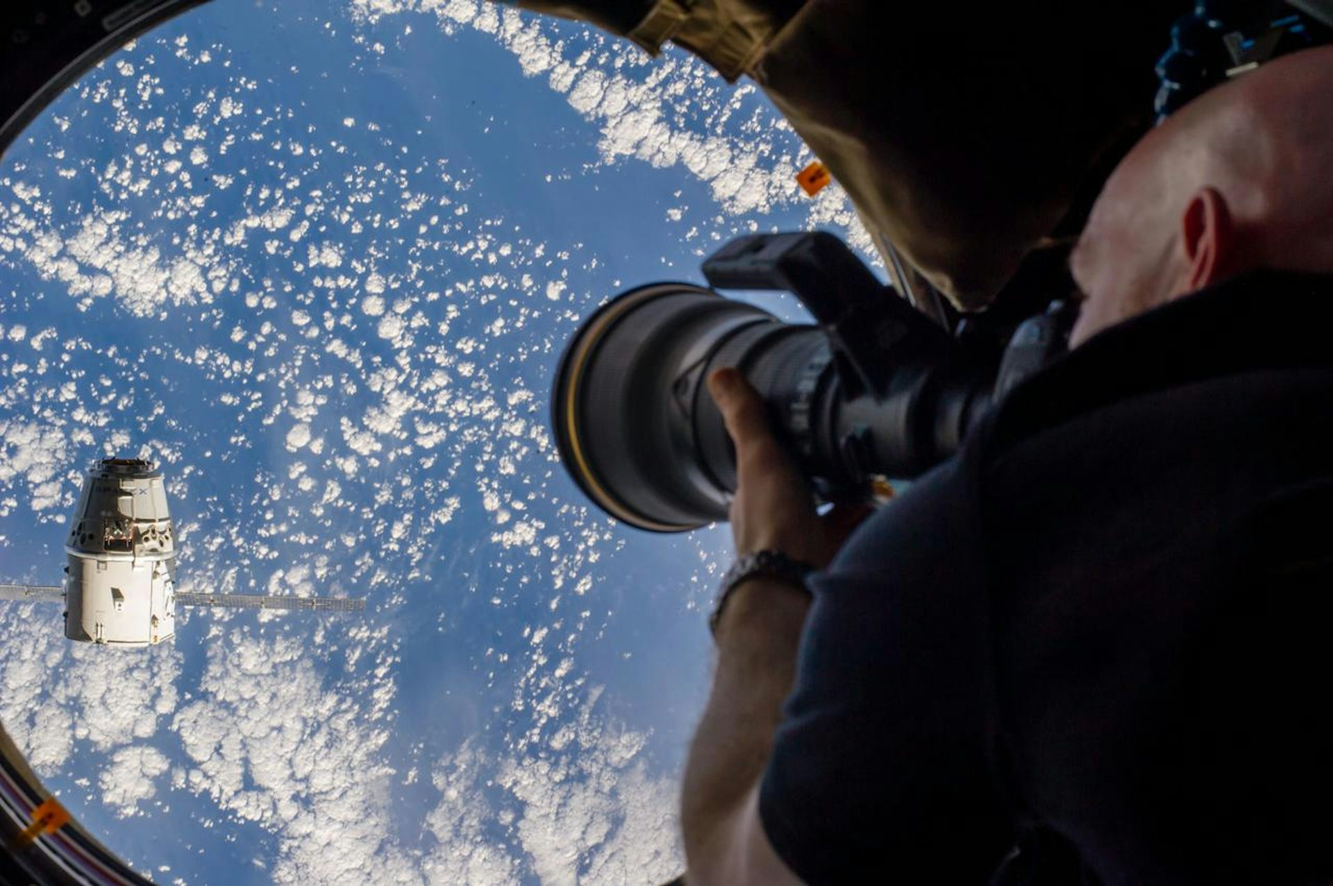 An astronaut pointing a camera out a circular window in the International Space Station at a