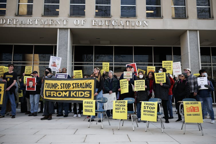 A large group of people hold yellow signs in front of a building. Some of the signs say