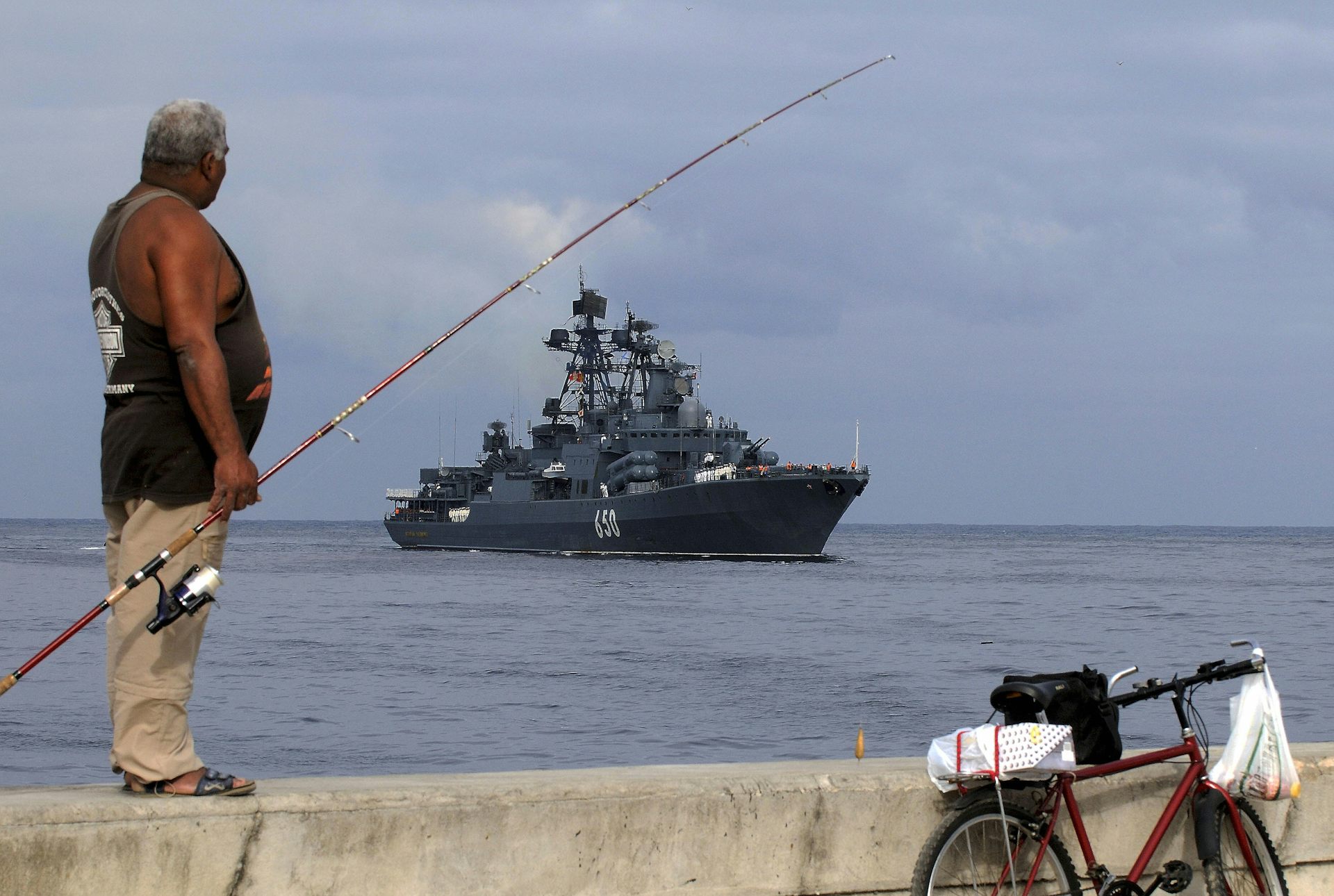 A man with a fishing rod looks out at sea where. large ship is.