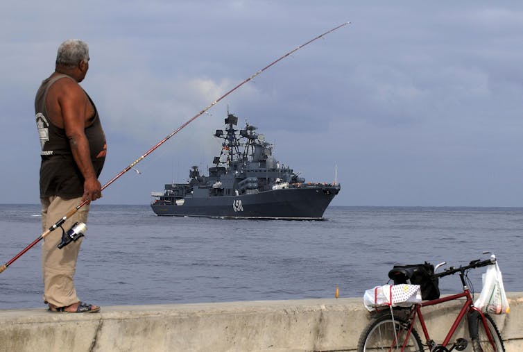 A man with a fishing rod looks out at sea where. large ship is.