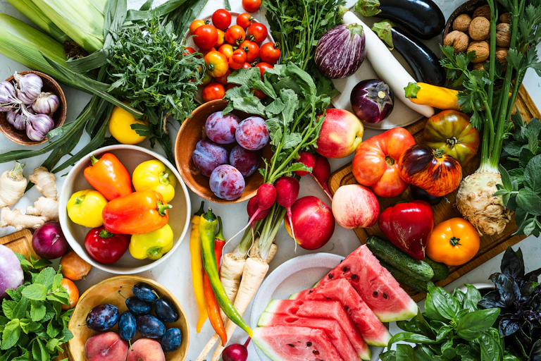 A wide variety of fruits and vegetables on a table, including watermelons, plums and tomatoes.