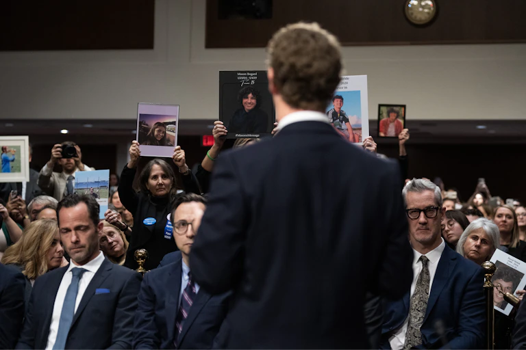 A man in a suit, Zuckerberg, is photographed from behind, standing in front of a gallery of people wearing somber expressions.