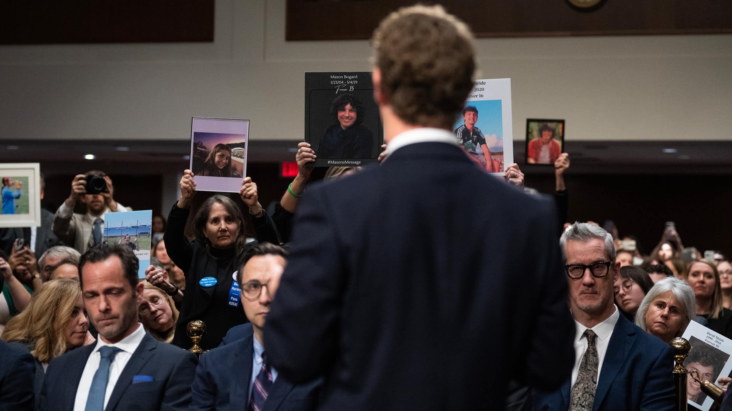 A man in a suit, Zuckerberg, is photographed from behind, standing in front of a gallery of people wearing somber expressions.