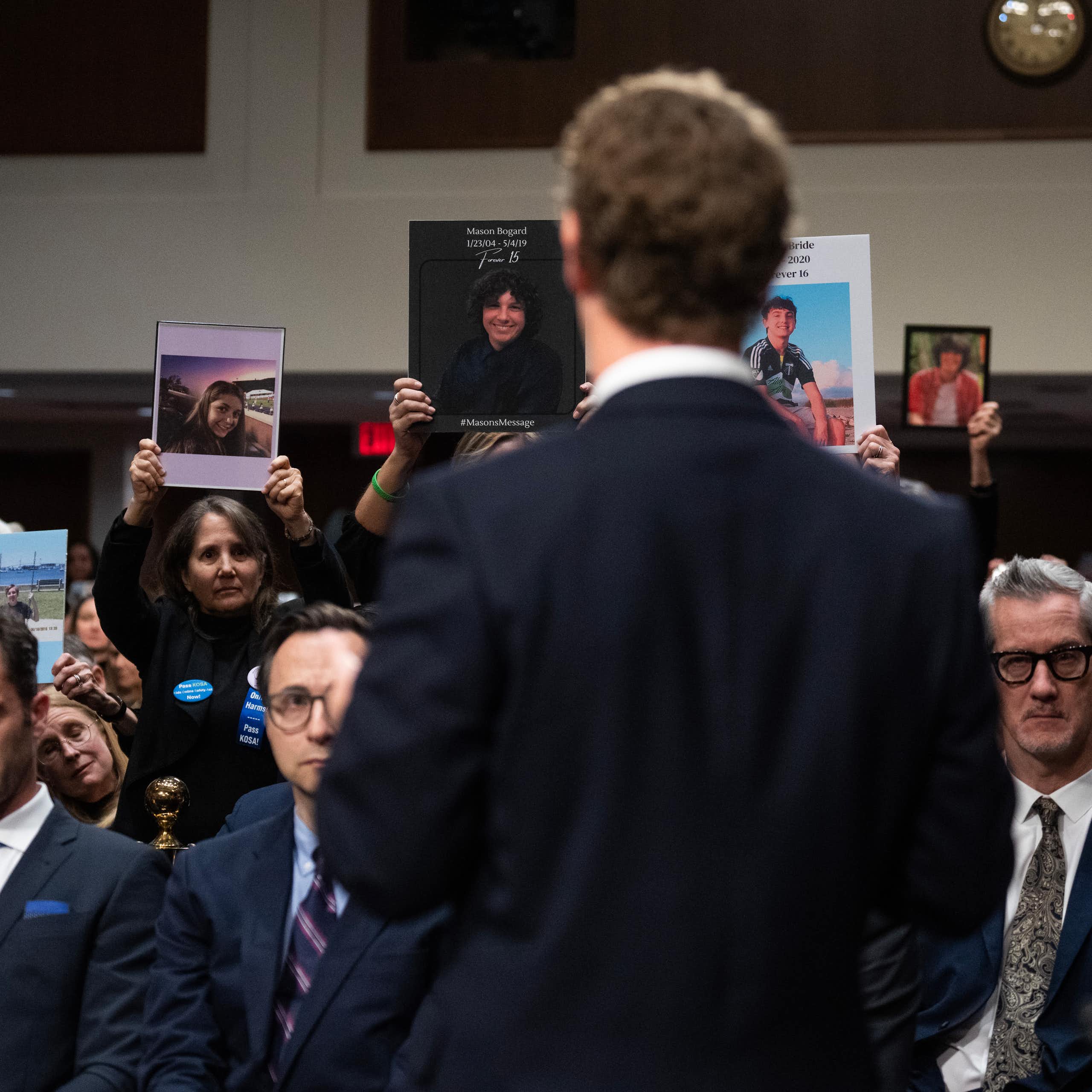 A man in a suit, Zuckerberg, is photographed from behind, standing in front of a gallery of people wearing somber expressions.