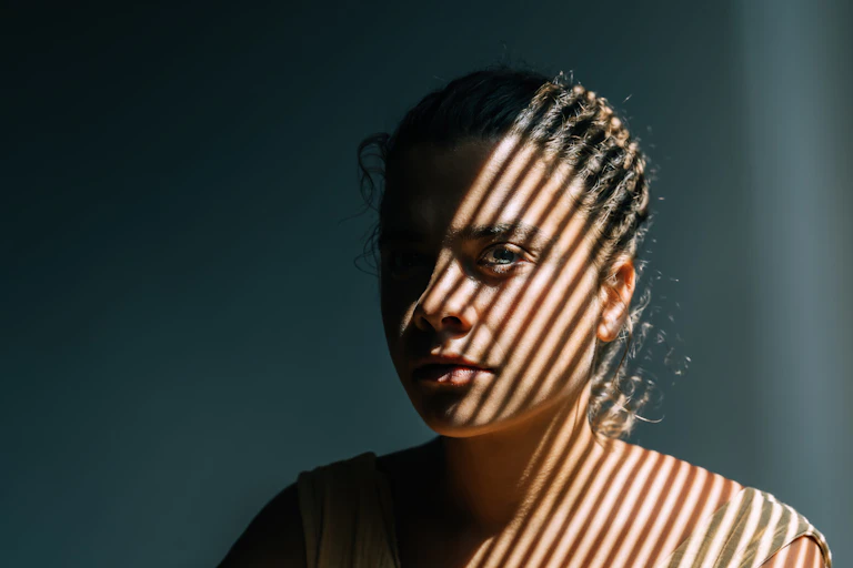 Woman sits next to a window in the striped light from a blind.