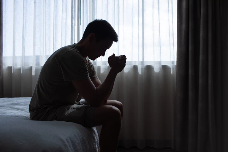 A young man sits on his bed in the dark. His hands are clasped together and he is looking thoughtful and serious.