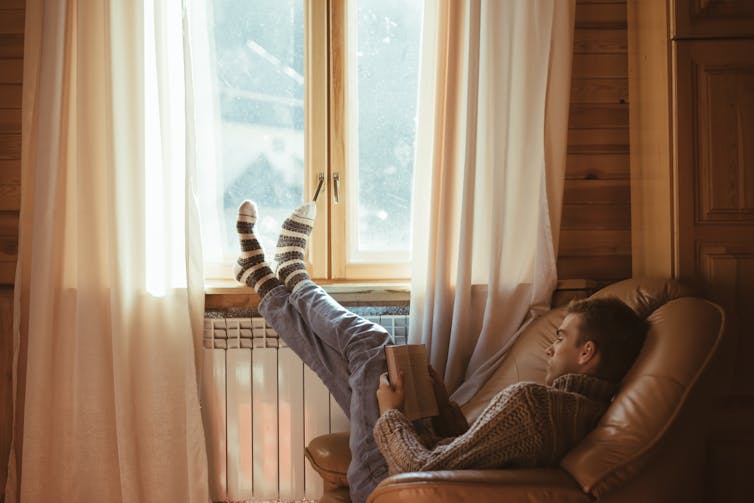 man sits on chair reading book by radiator, keeping warm inside