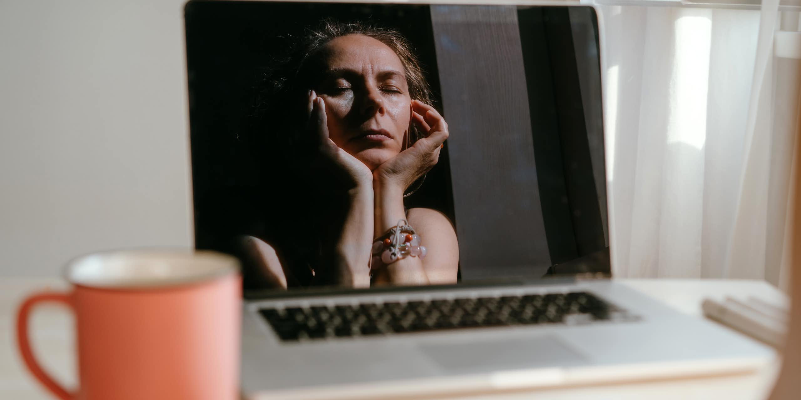 woman's reflection in laptop shows her closing her eyes and massaging her temples