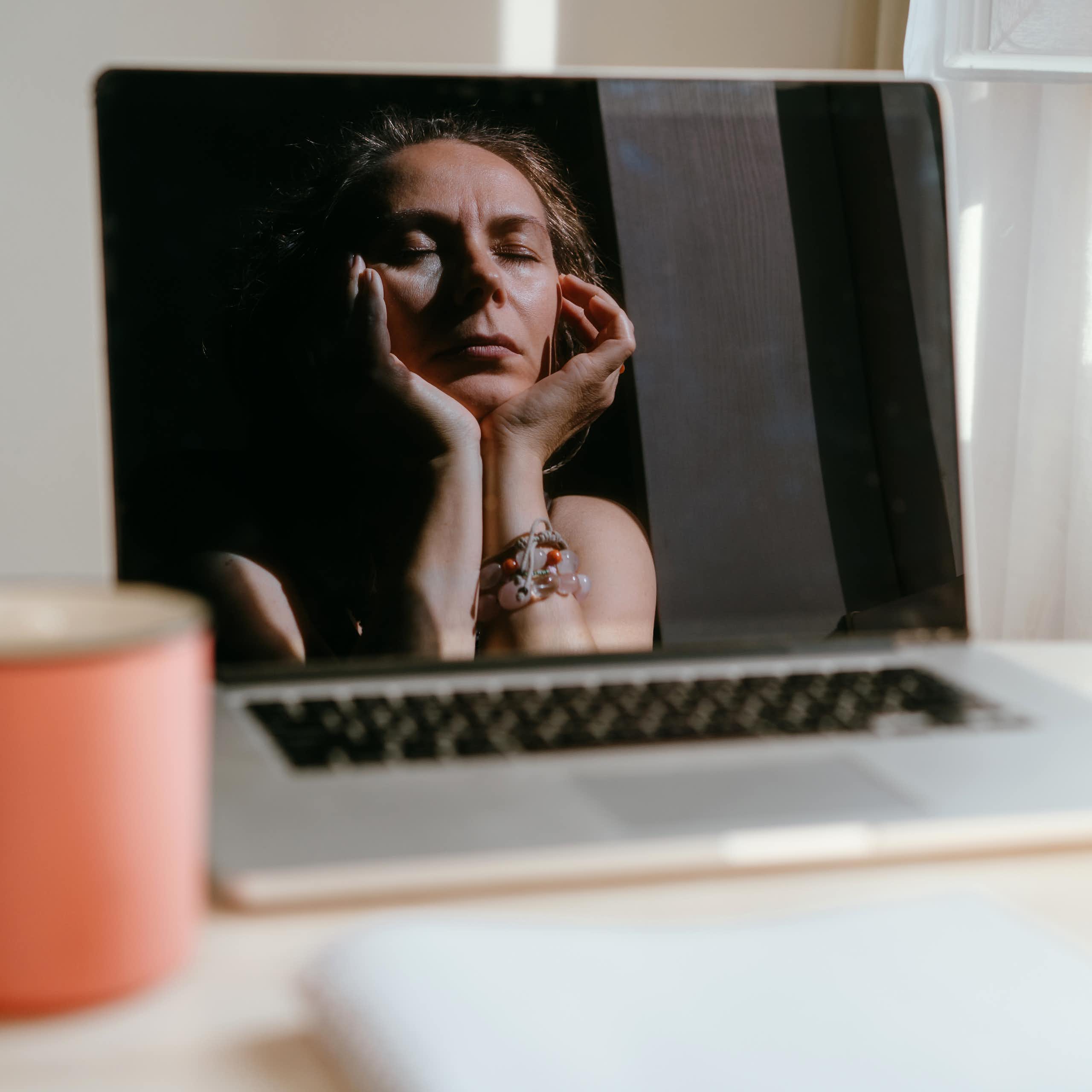 woman's reflection in laptop shows her closing her eyes and massaging her temples