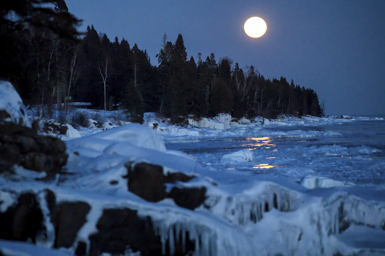 rive gelée d'un lac bordée d'arbres. Une pleine lune dans le ciel au-dessus.