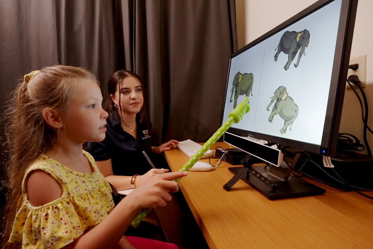 A young girl with blonde hair and a yellow shirt holds a green plastic pointer and looks at a screen that has cartoon elephants on it. A woman with dark hair sits next to her.