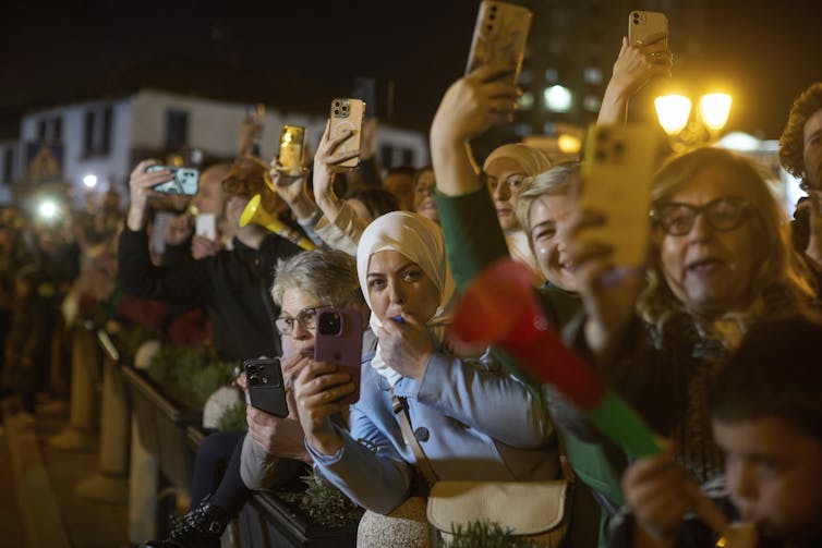 Camera-wielding people wait expectantly behind a line for the arrival of others.