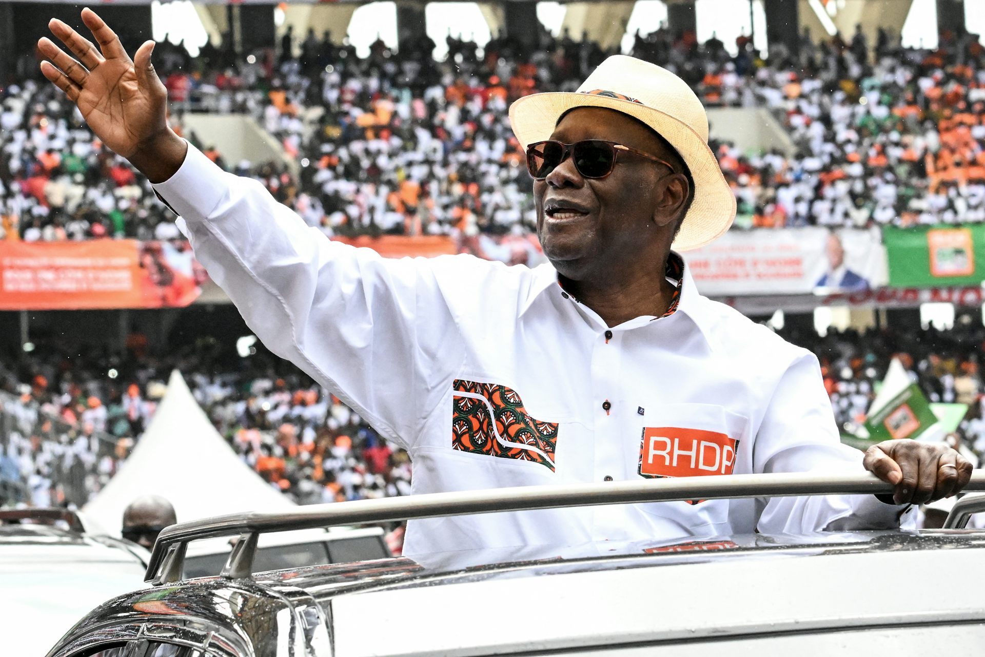 Man wearing a white shirt and bowler hat, waving at a large crowd 