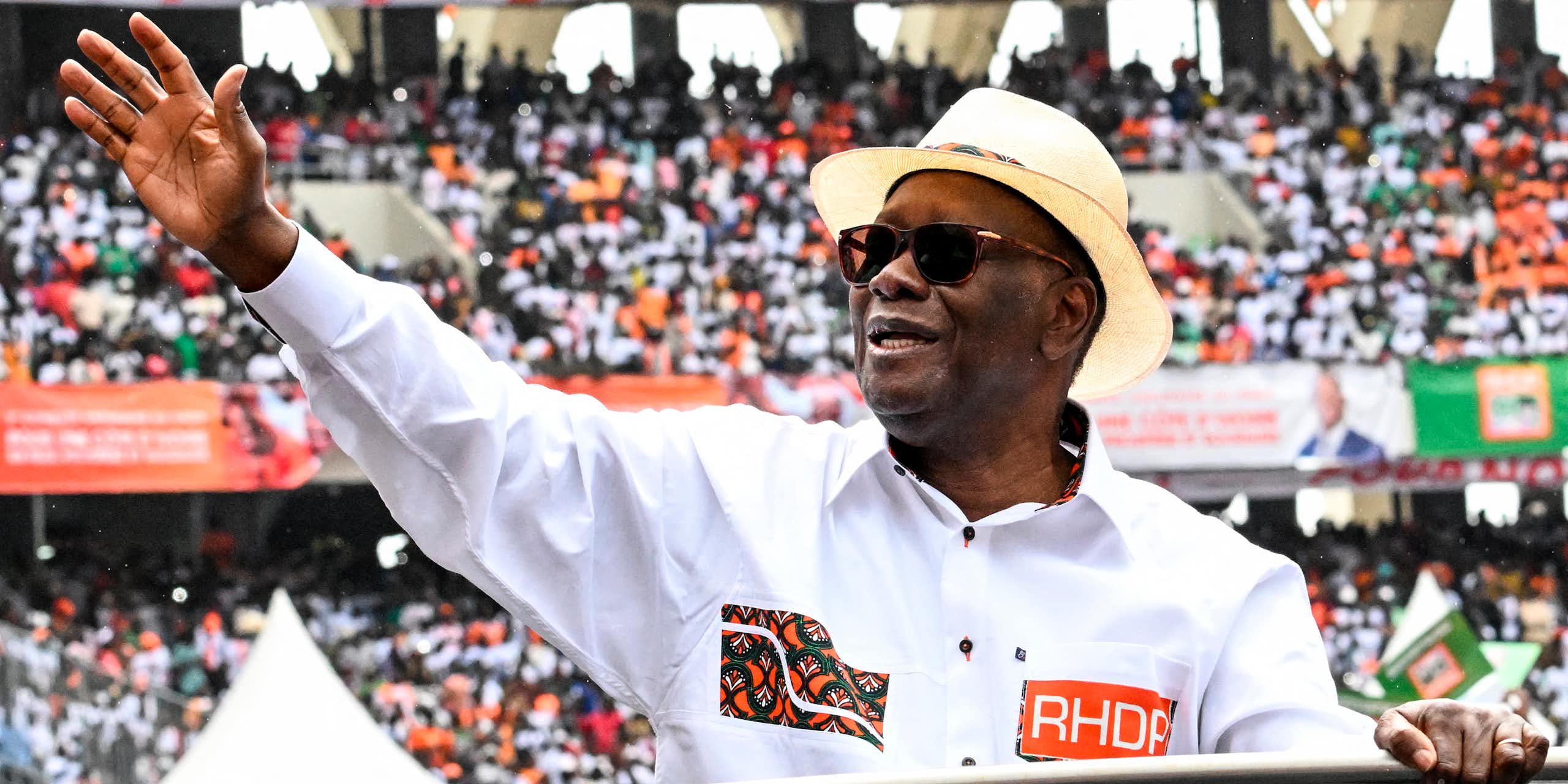 Man wearing a white shirt and bowler hat, waving at a large crowd