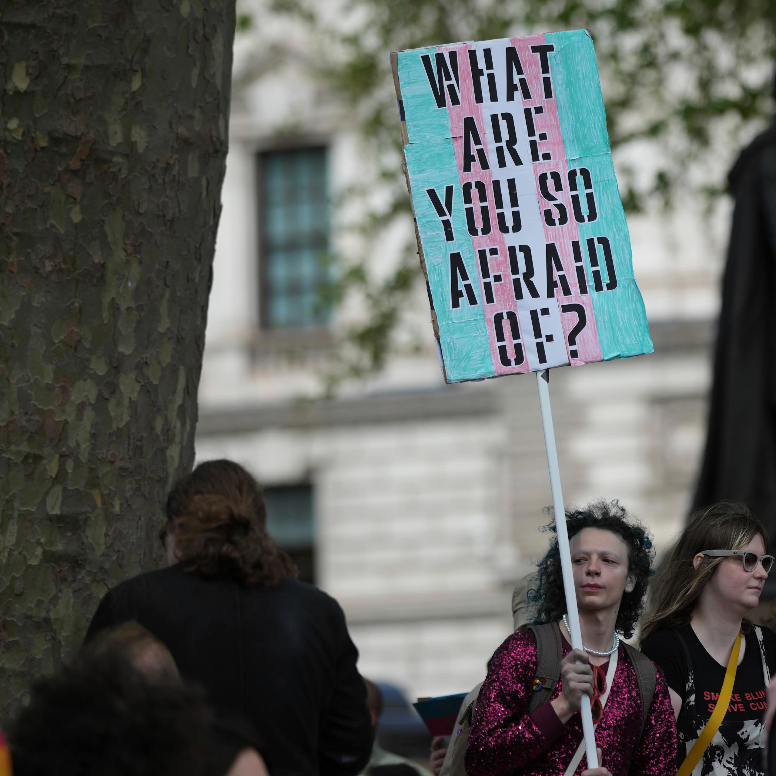Protestor holding a sign reading 'WHAT ARE YOU SO AFRAID OF?' painted in the trans flag colors