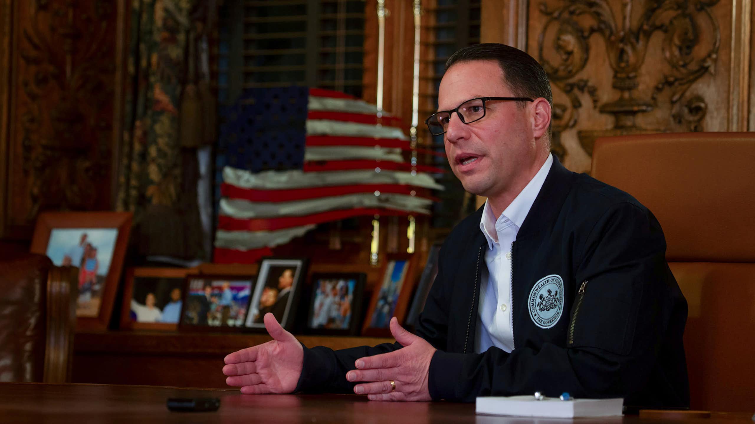 Man in suit jacket and white shirt and wearing glasses sits at table with American flag in background